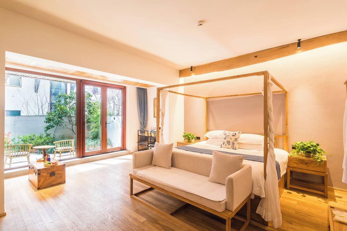 A bedroom with a canopy bed, a sofa, a small table, and a view of the outdoors through large windows.
