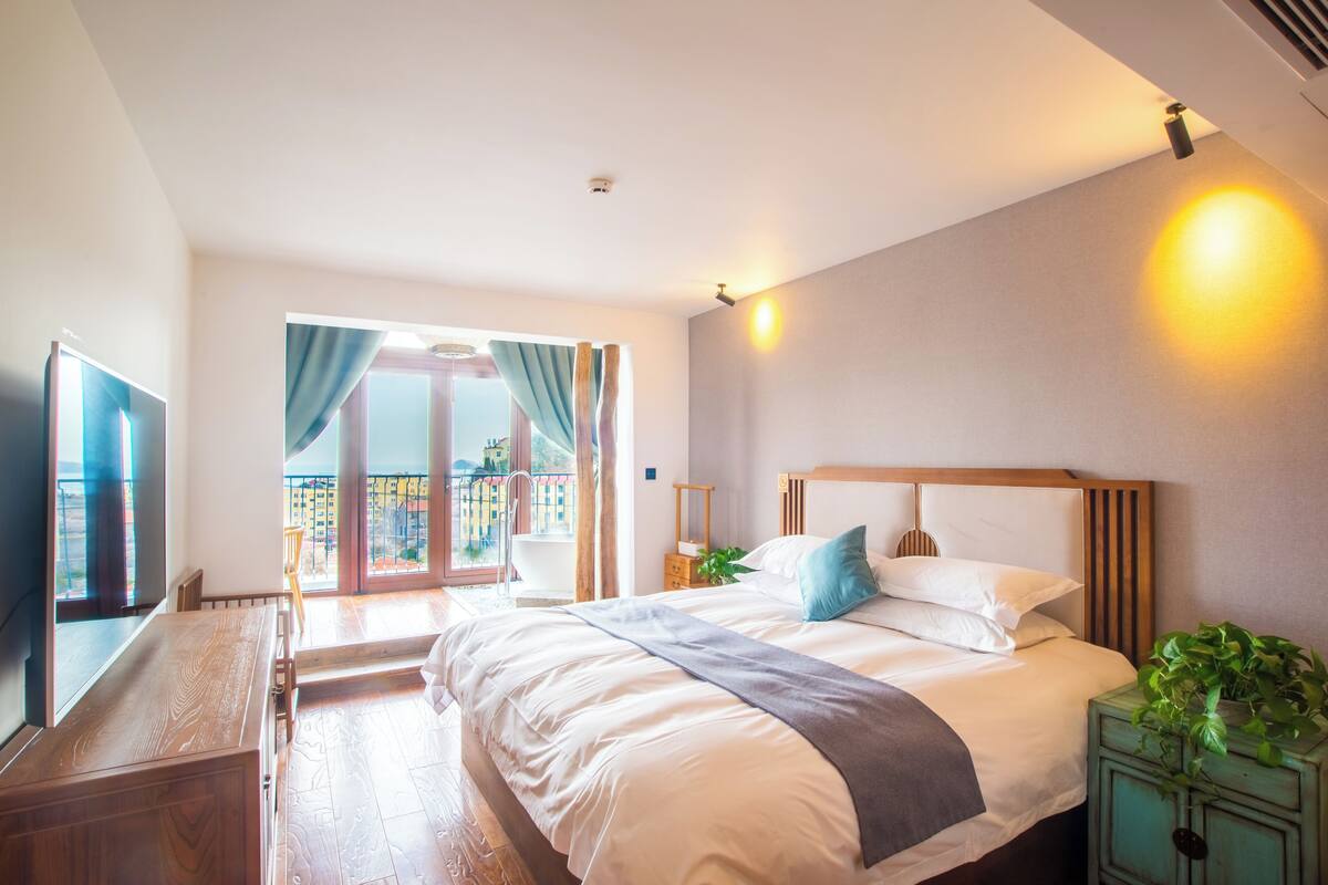 A bedroom with a large bed, a wooden nightstand, a TV, and a balcony with curtains.