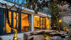 Una casa moderna con grandes puertas de vidrio, un muro de piedra y un pequeño estanque rodeado de rocas.