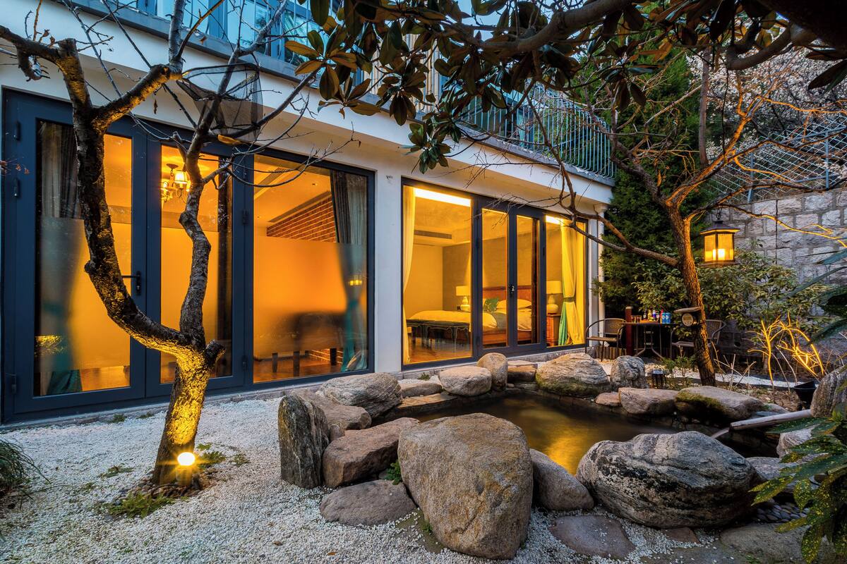 A modern house with large glass doors, a stone wall, and a small pond surrounded by rocks.