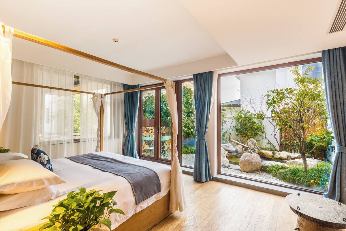 A bedroom with a large bed, a view of a garden through floor-to-ceiling windows, and a small round table.