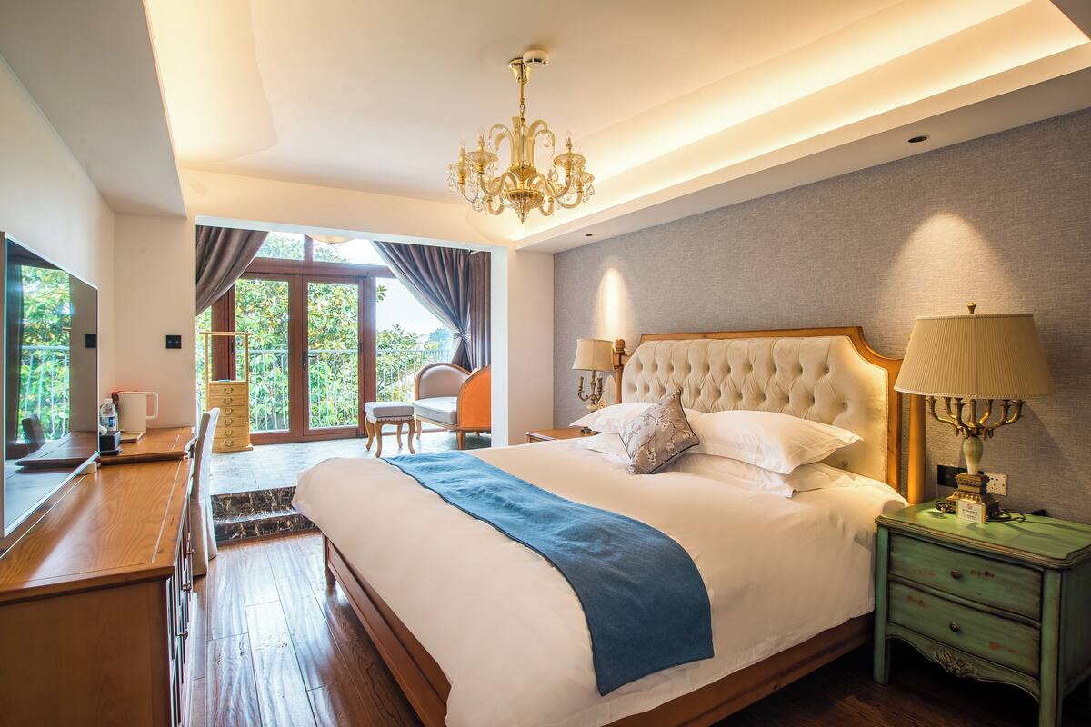 A spacious bedroom with a large bed, a wooden desk, a chair, and a view of greenery outside.