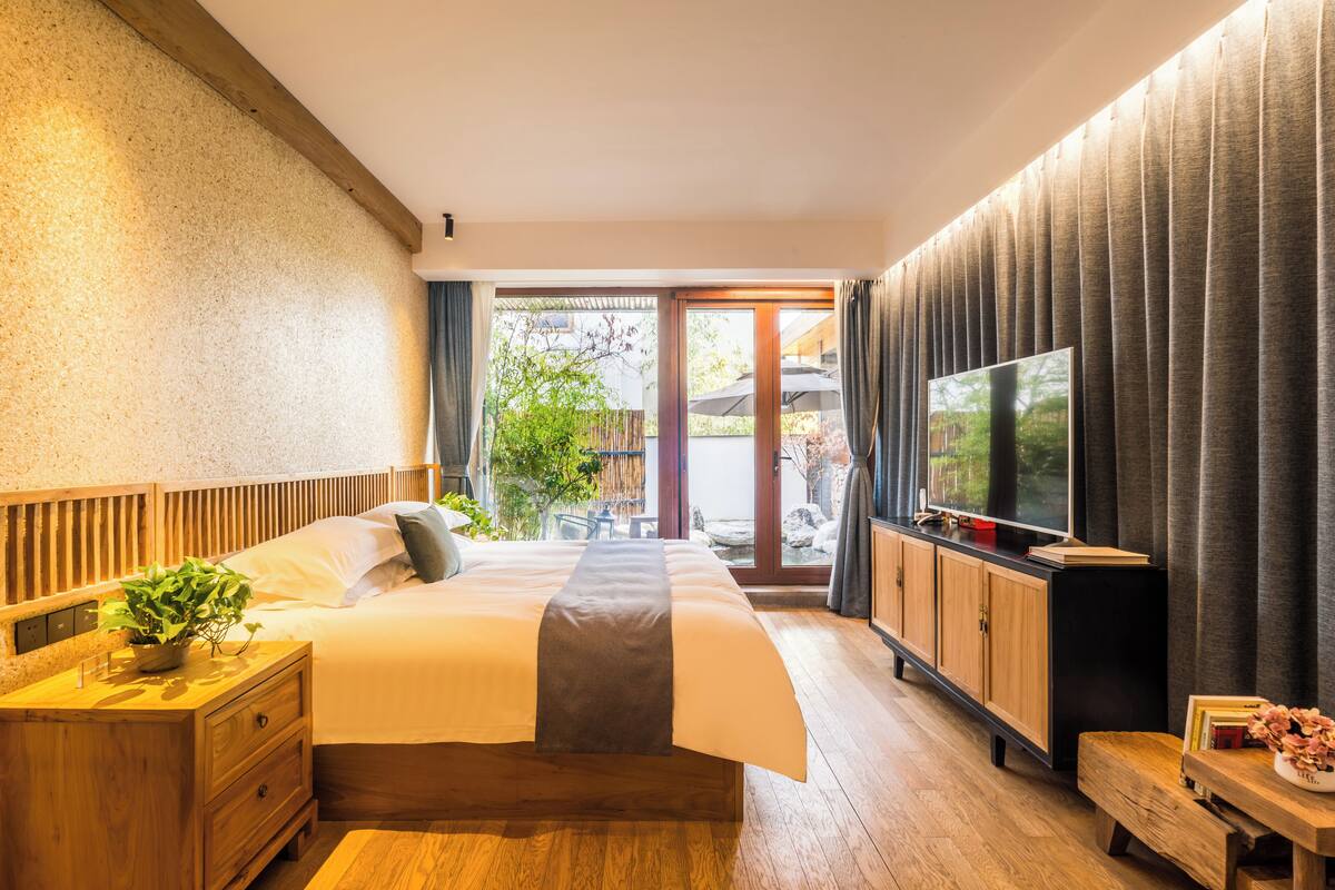 A modern hotel room with a large bed, wooden headboard, a TV, and a view of the outdoors.