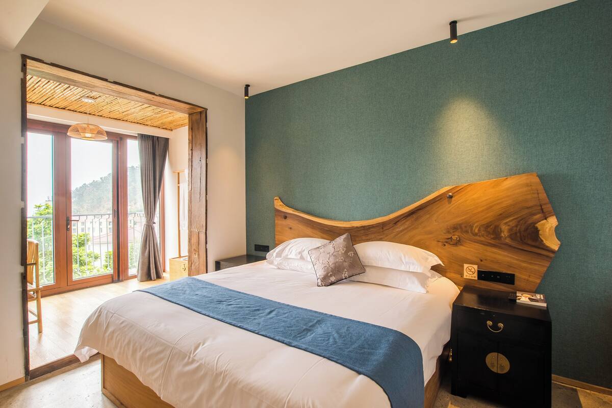 A bedroom with a large bed, a wooden headboard, a nightstand, and a view through a sliding door to an outdoor balcony.