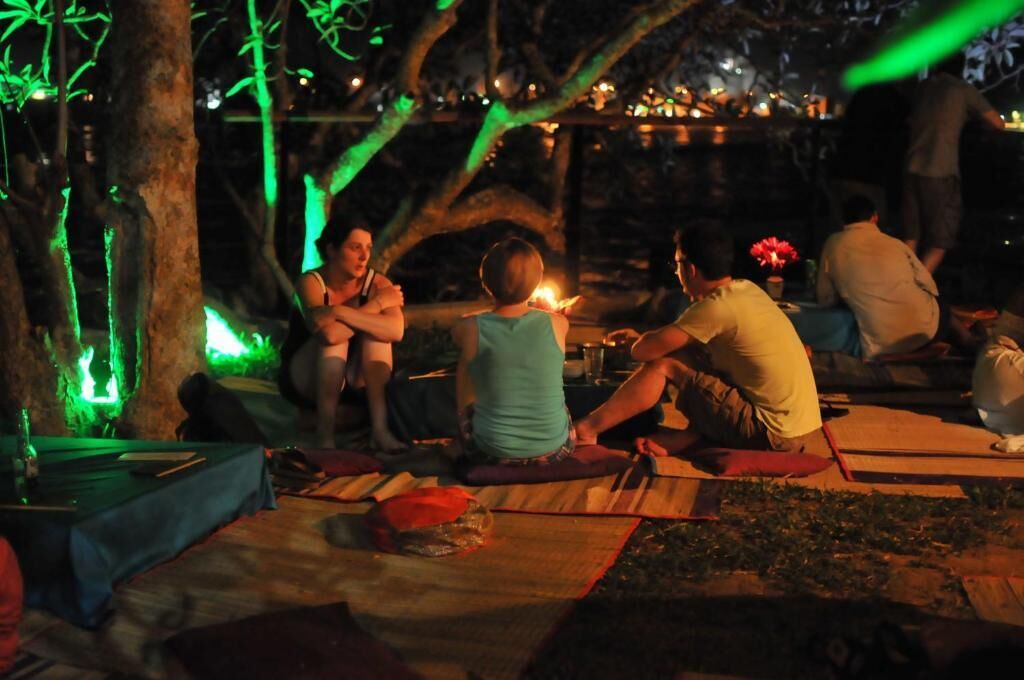 Een groep mensen zit 's avonds buiten, mogelijk op een camping of evenement, met een tafel ervoor.