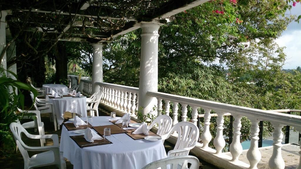 A covered outdoor dining area with white chairs and tables, surrounded by greenery and a white balustrade.