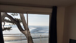 Ein Zimmer mit einer großen Schiebeglastür, die auf einen Strand blickt, einem kleinen runden Tisch, einem Holzstuhl und einem Fernsehschrank.
