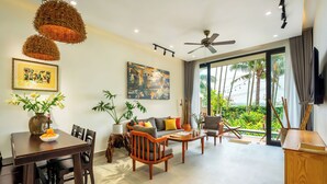A modern living room with a sofa, coffee table, and dining area. There is a ceiling fan, large windows, and a view of palm trees.