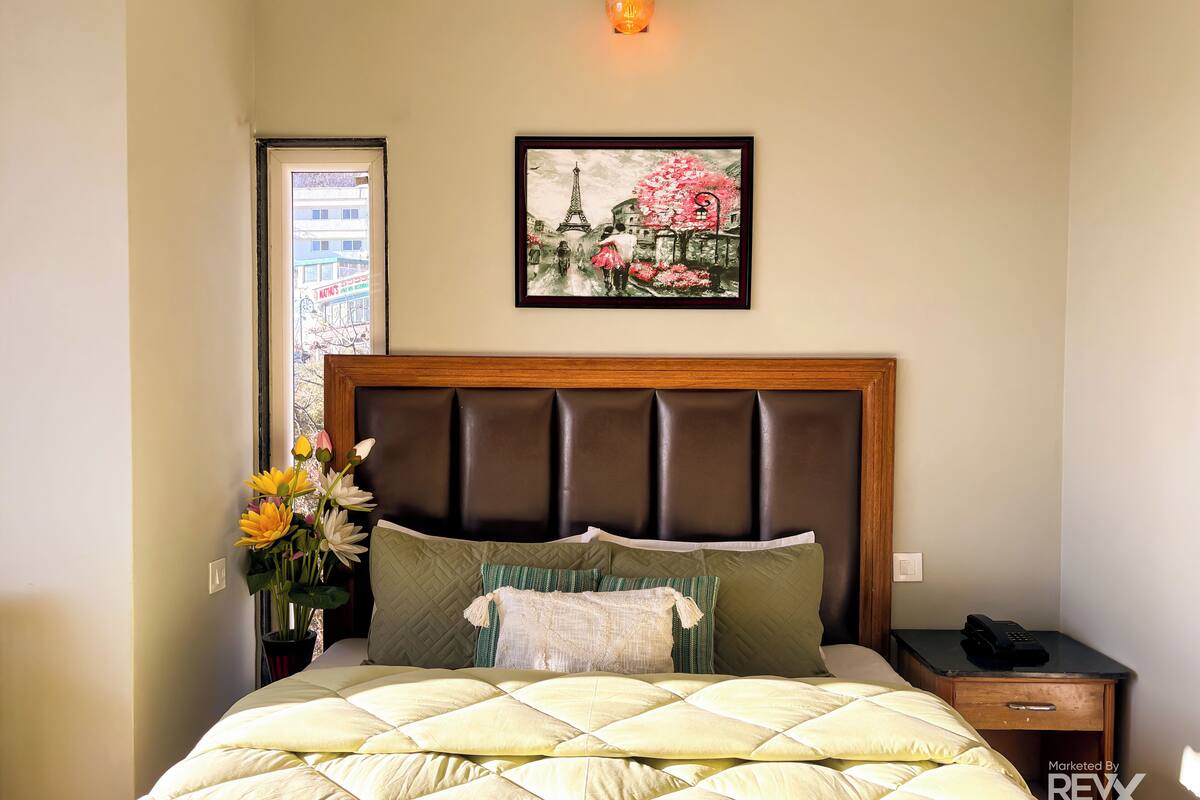 A neatly made bed with a quilted headboard, a bedside table with a lamp, and a framed picture of the Eiffel Tower on the wall.
