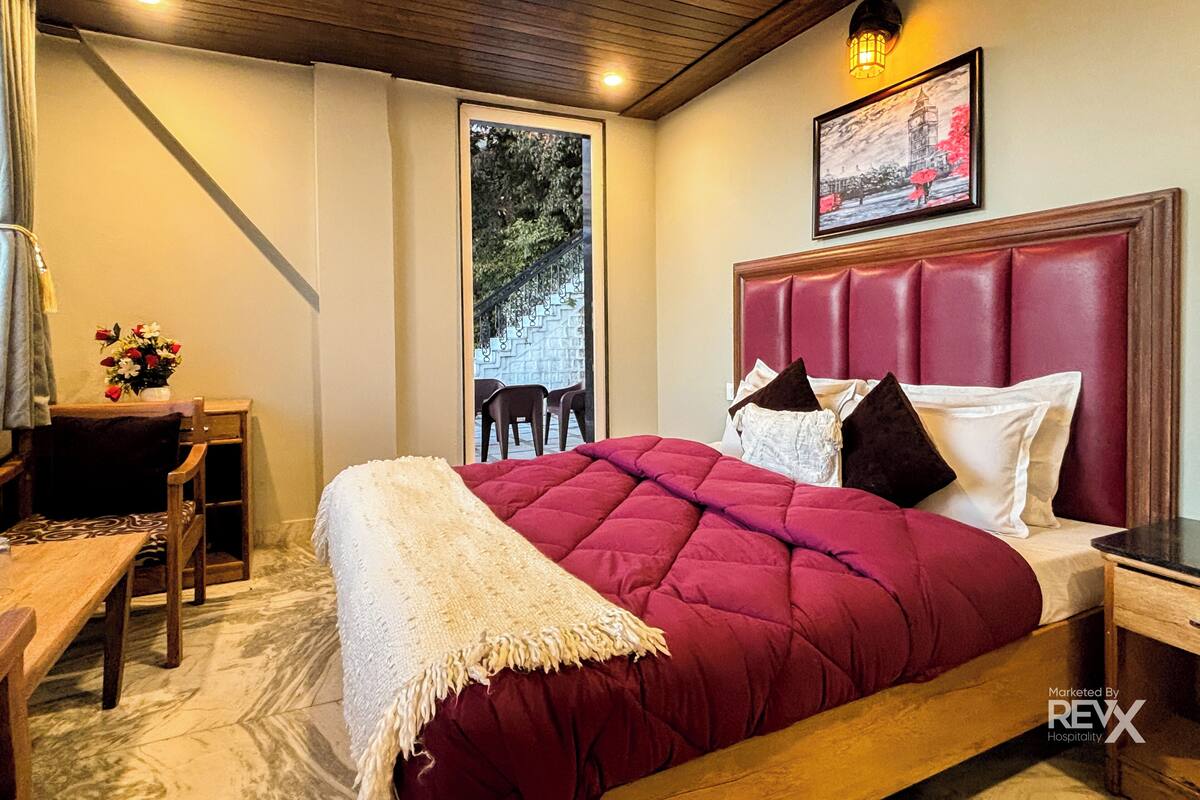 A hotel room with a large bed, a wooden headboard, a nightstand, a desk with a chair, and a window with a view of a staircase and greenery.
