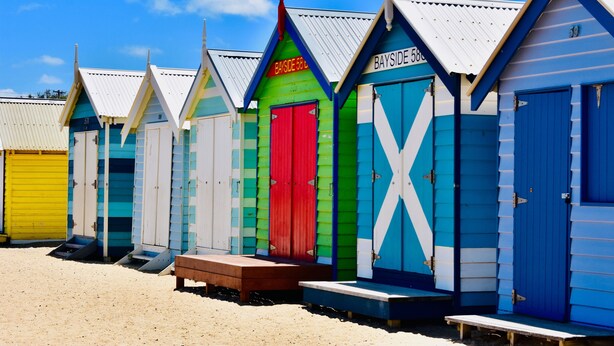 Une rangée de cabanes de plage colorées, chacune dotée d’une porte numérotée et d’un petit banc devant.