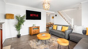 Un salon moderne avec un canapé gris, des coussins jaunes, un pouf rond orange, un téléviseur à écran plat et un escalier menant à un autre niveau.