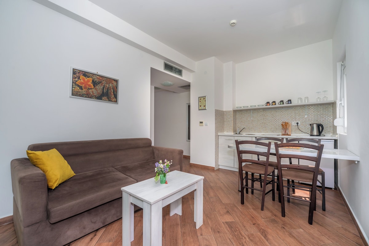 A compact living space with a brown sofa, a small white table, and a dining area with wooden chairs. A kitchen with white cabinets and a tiled backsplash is visible in the background.