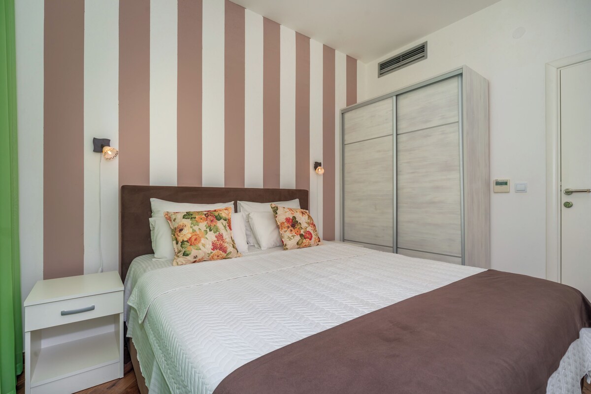 A bedroom with a bed, a nightstand, a wardrobe, and a wall with vertical stripes.