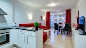A compact kitchen with white cabinets and a black countertop, a red sofa, a dining table with chairs, and a TV on a white stand.
