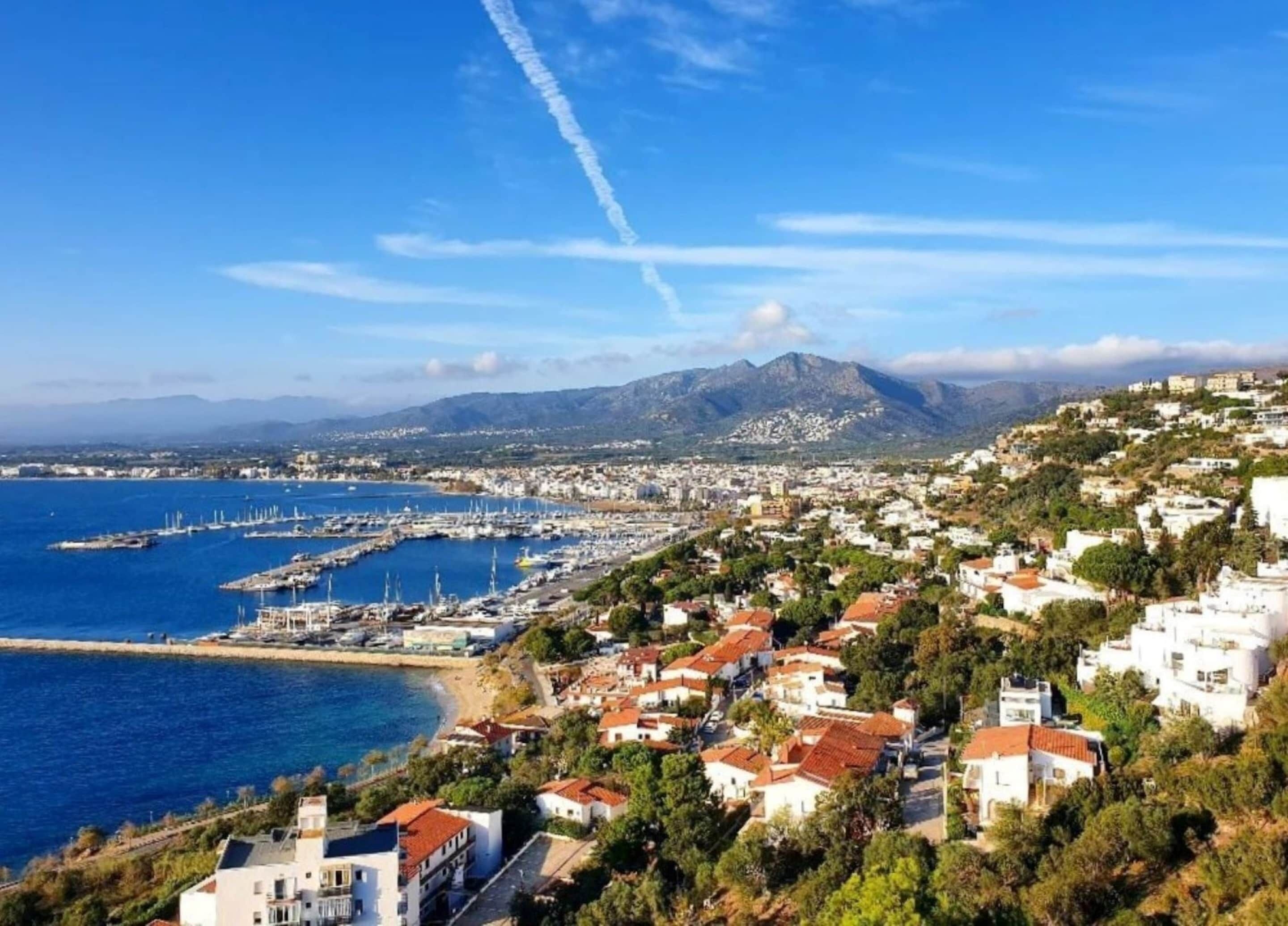 Un poble costaner amb un port esportiu, edificis blancs i una muntanya al fons.