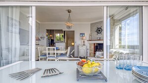 Un coin repas avec une table dressée pour deux personnes, un panier de fruits et une vue sur l’intérieur à travers une porte vitrée.