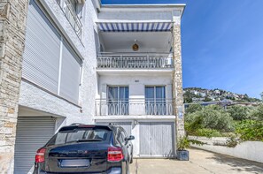 Un immeuble à plusieurs étages avec une voiture garée devant, un balcon doté d’une balustrade et une vue sur une colline parsemée de maisons.