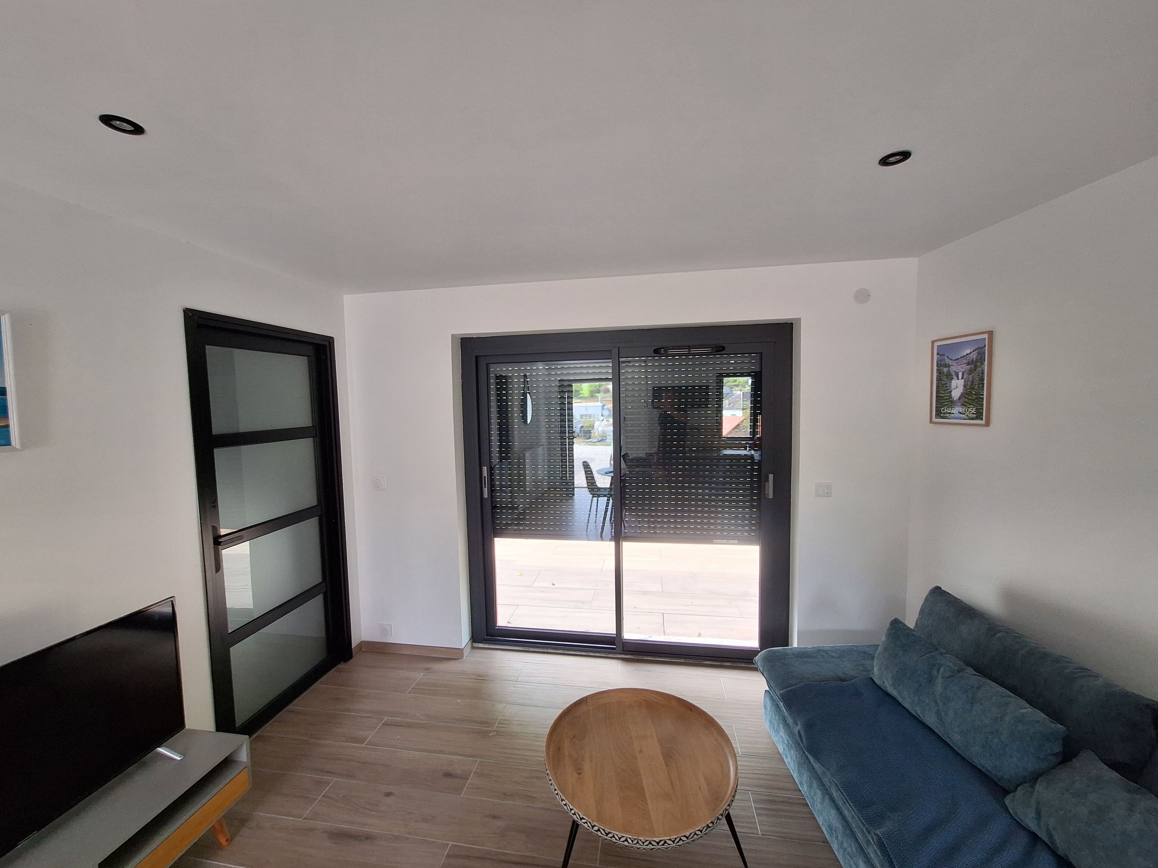 Un salon moderne comprenant un téléviseur, un canapé, une chaise en bois et une porte-fenêtre coulissante donnant sur un espace extérieur.