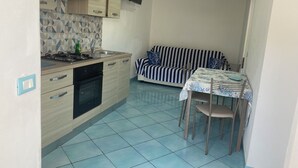 A compact kitchen with a gas stove, oven, and sink, a striped sofa, and a small dining table with chairs. - Ponzamania Sea urchin (Le Forna)