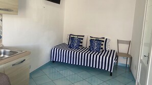 A small, compact room with a bed, a chair, a TV mounted on the wall, and a kitchenette area. - Ponzamania Sea urchin (Le Forna)