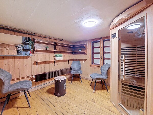 Une pièce chaleureuse avec des murs en bois, un radiateur, deux chaises, une petite poubelle et une fenêtre avec des stores.
