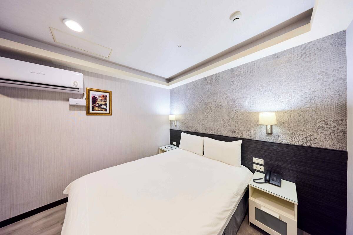 A hotel room with a bed, bedside table with a phone, wall-mounted air conditioning unit, and a picture on the wall.