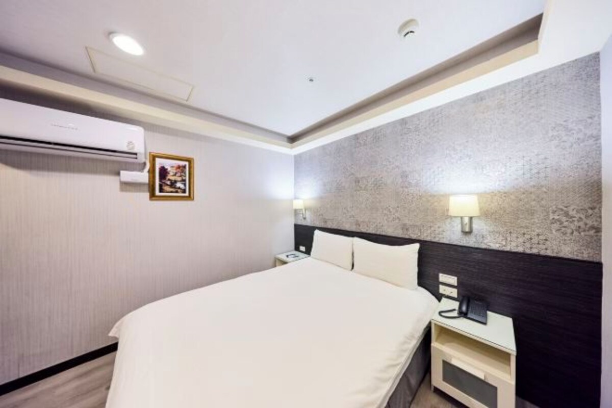 A hotel room with a bed, bedside table, nightstand, wall-mounted air conditioning unit, and a textured wall.
