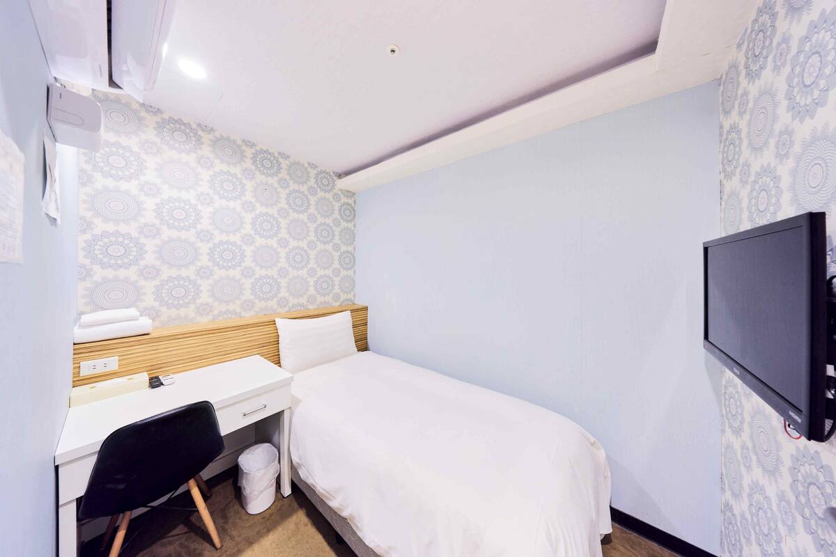 A hotel room with a bed, a desk with a chair, a television, and patterned wallpaper.