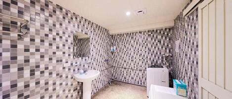 A bathroom with a tiled wall, a sink, a mirror, and a shower area.