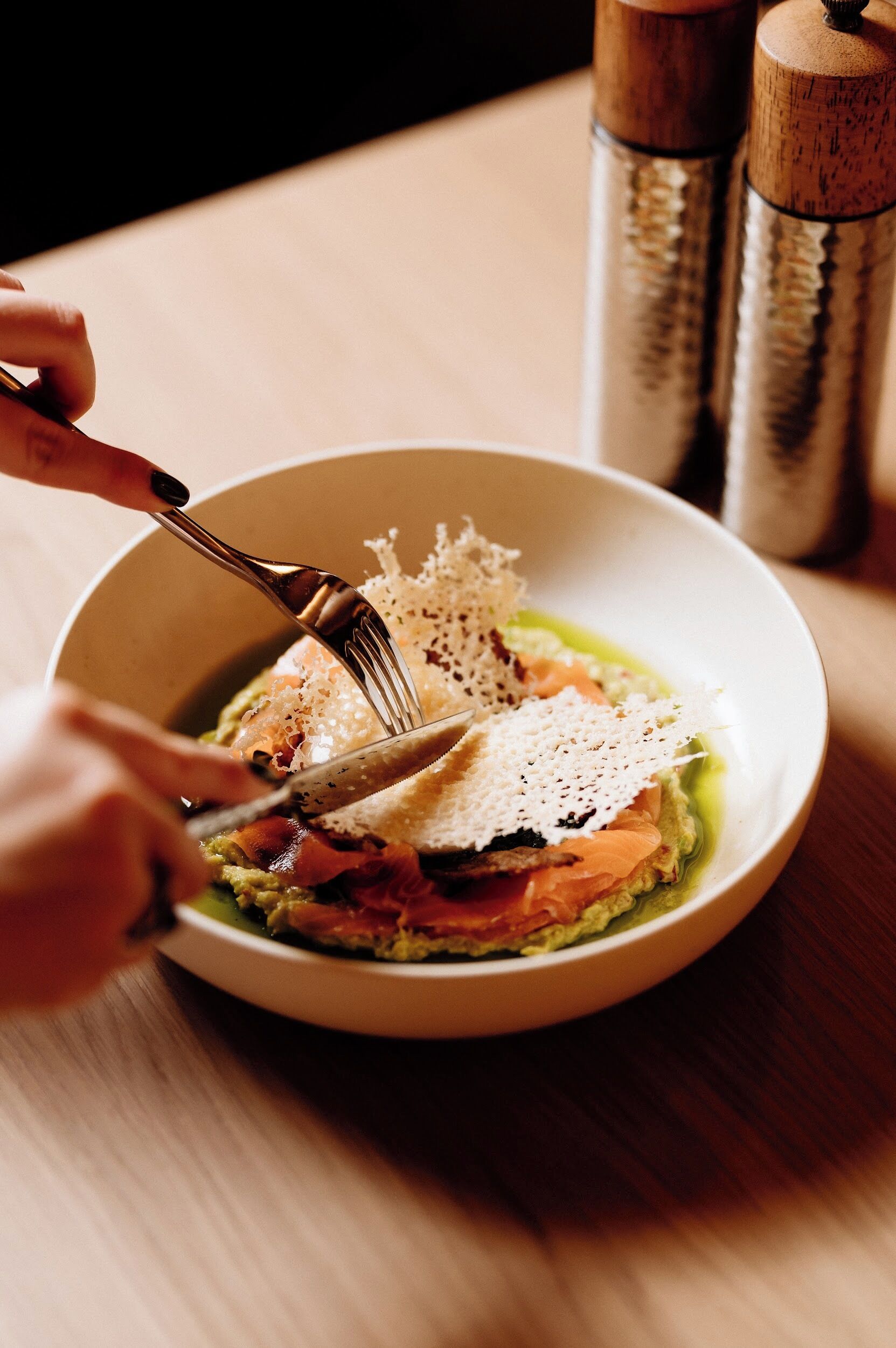 A person using a fork and knife to eat a dish with salmon and greens.