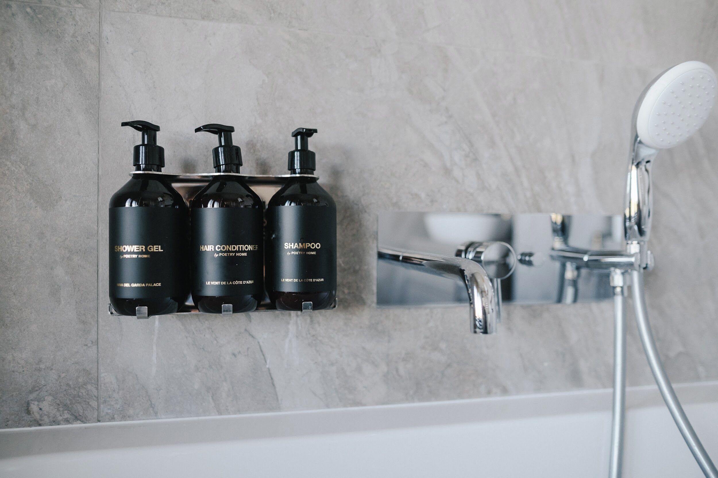 Un baño con pared de mármol, un estante con tres botellas negras etiquetadas como “Gel de Ducha”, “Acondicionador para el Cabello” y “Champú”, y un cabezal de ducha moderno.