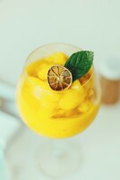 A cocktail glass with a yellow drink, garnished with a dried orange slice and a mint leaf.