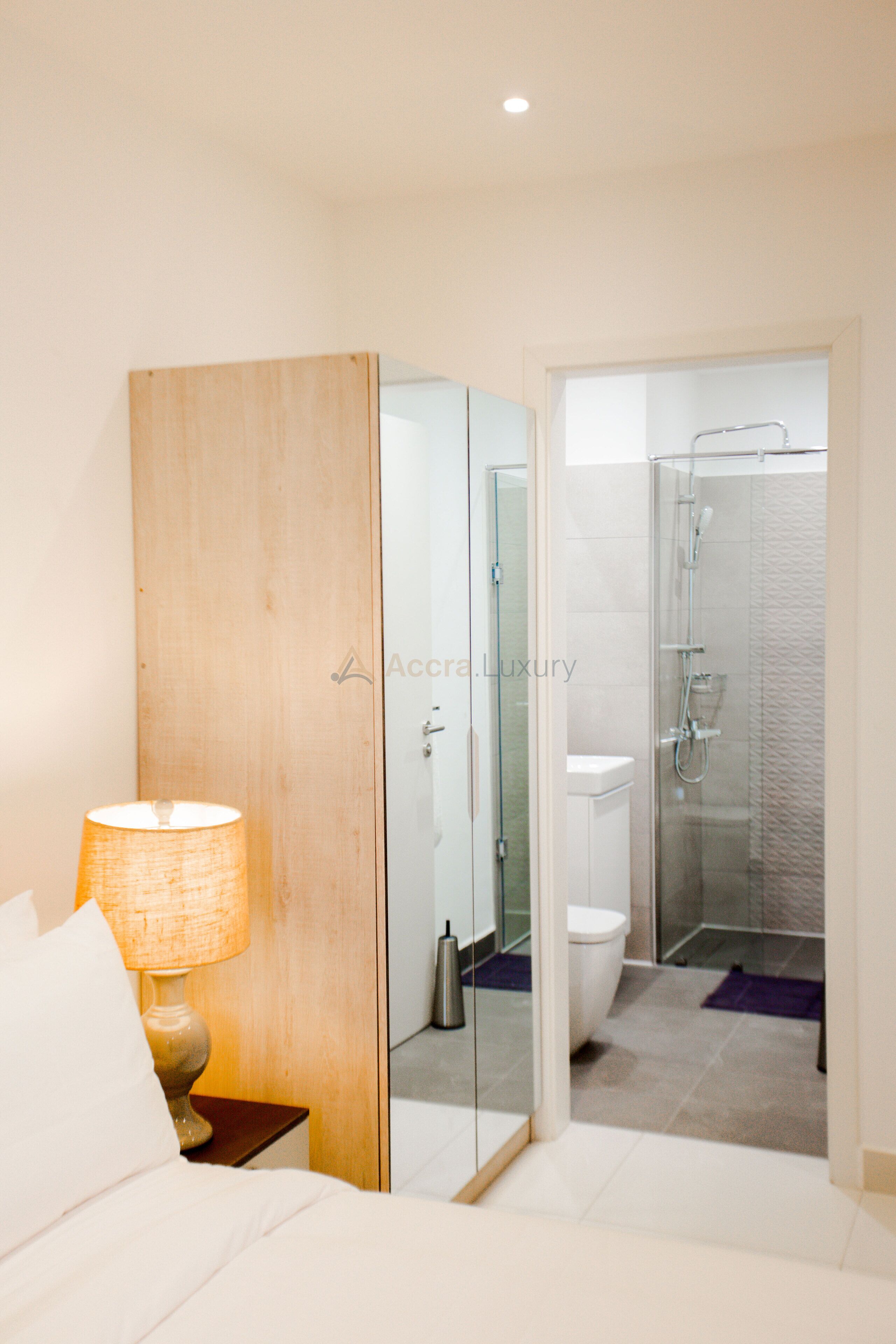 A hotel room with a bed, a nightstand with a lamp, a wooden wardrobe, a glass partition, a toilet, and a shower area.