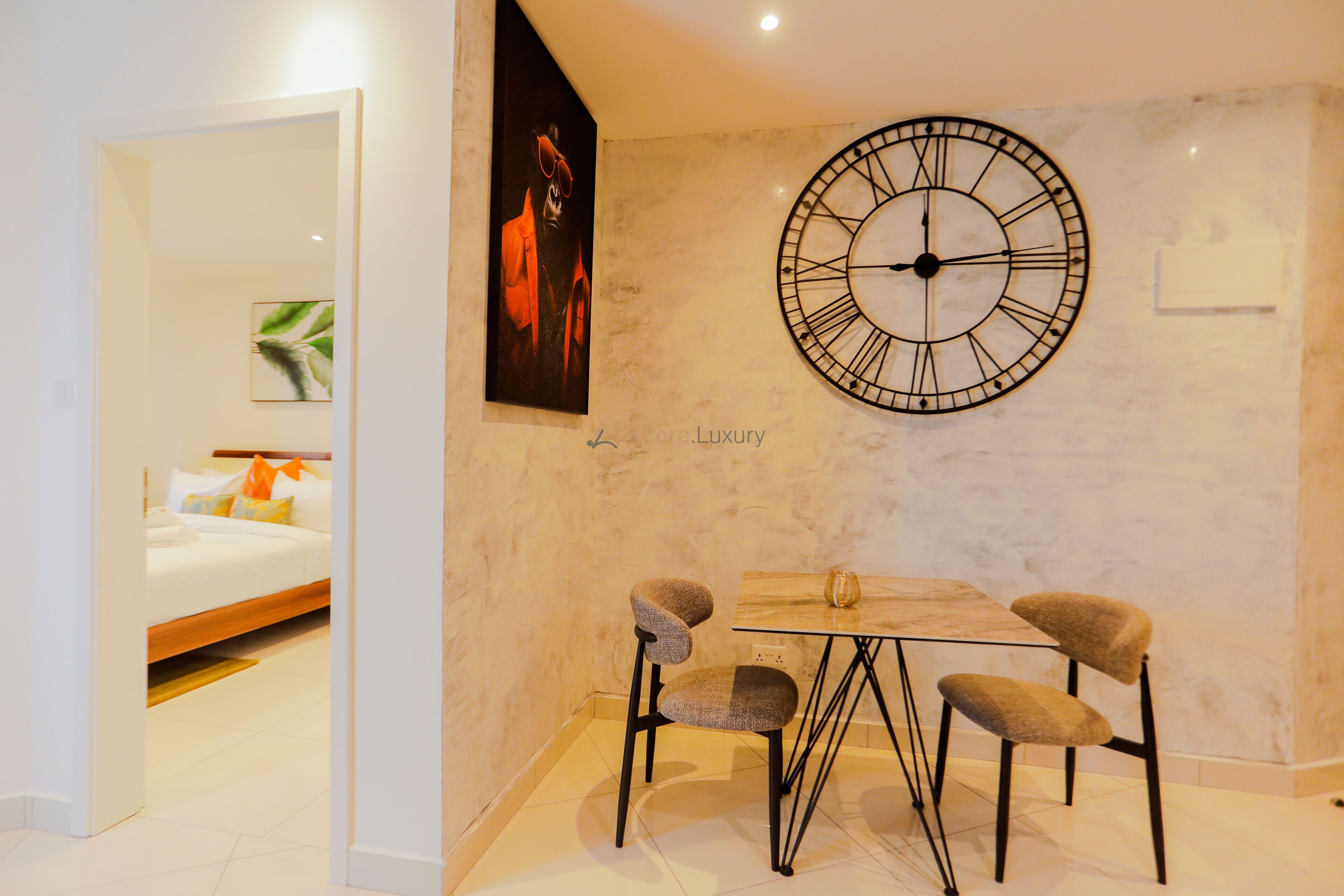 A modern interior with a large wall clock, a dining table with four chairs, and a bed with pillows in the background.