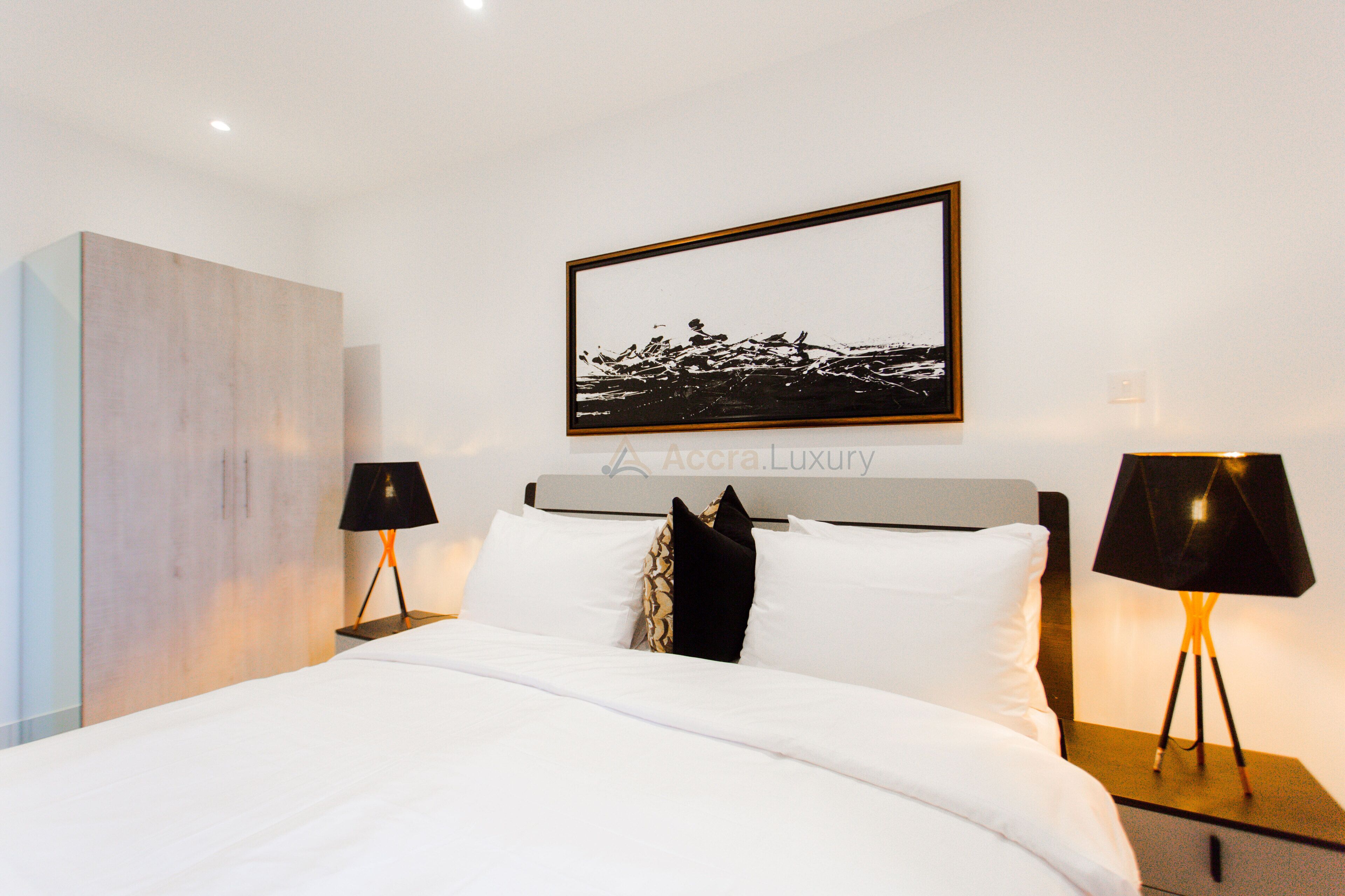 A modern hotel room with a large bed, two bedside tables with lamps, a framed artwork above the bed, and a minimalist wall design.