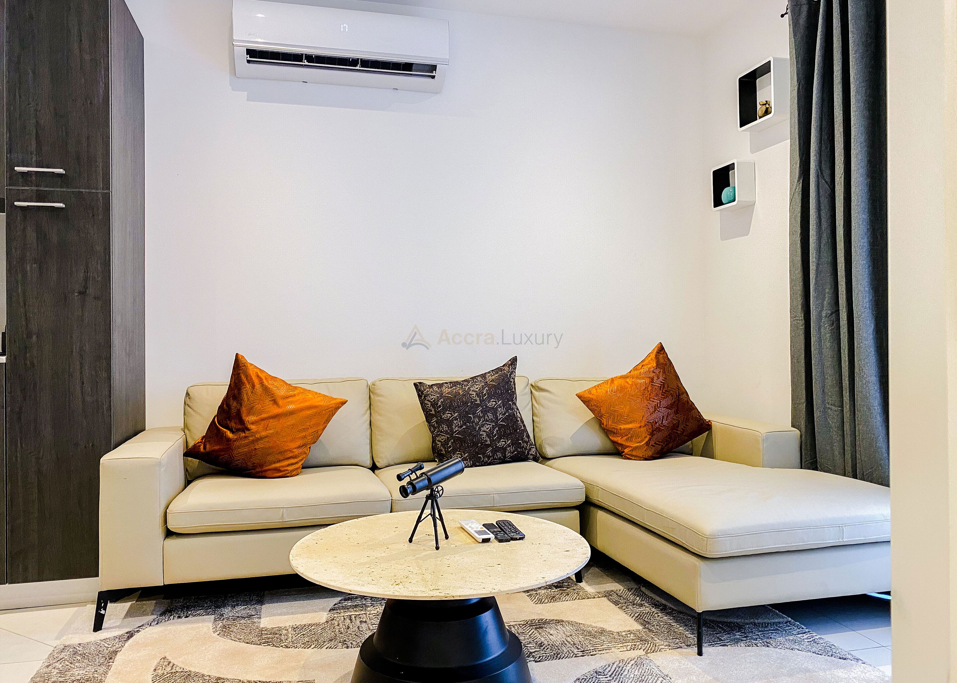 A modern living room with a beige sectional sofa, two orange and one black patterned pillow, a small round coffee table with a telescope and remote controls, and a wall-mounted air conditioner.
