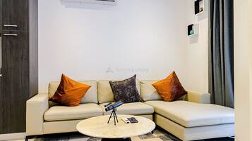 A modern living room with a beige sectional sofa, two orange and one black patterned pillow, a small round coffee table with a telescope and remote controls, and a wall-mounted air conditioner.
