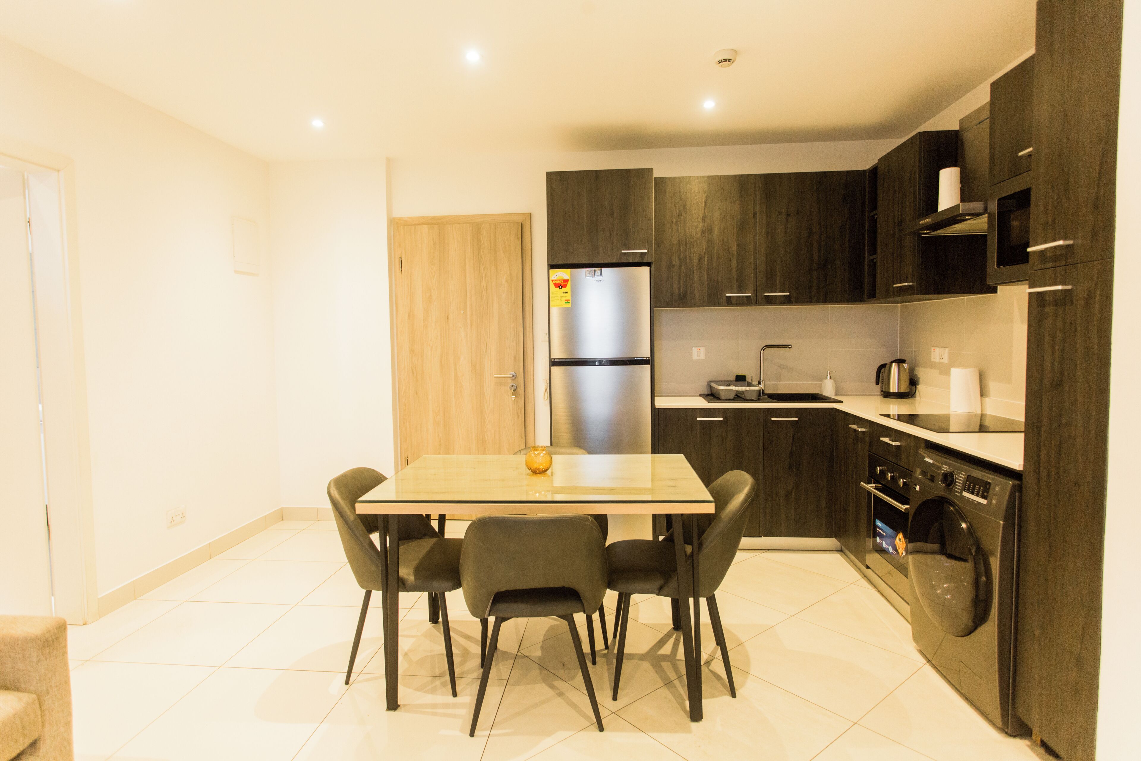 A modern kitchen with dark cabinetry, a stainless steel refrigerator, a built-in oven, and a dining area with a table and chairs.