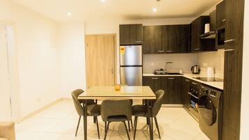 A modern kitchen with dark cabinetry, a stainless steel refrigerator, a built-in oven, and a dining area with a table and chairs.