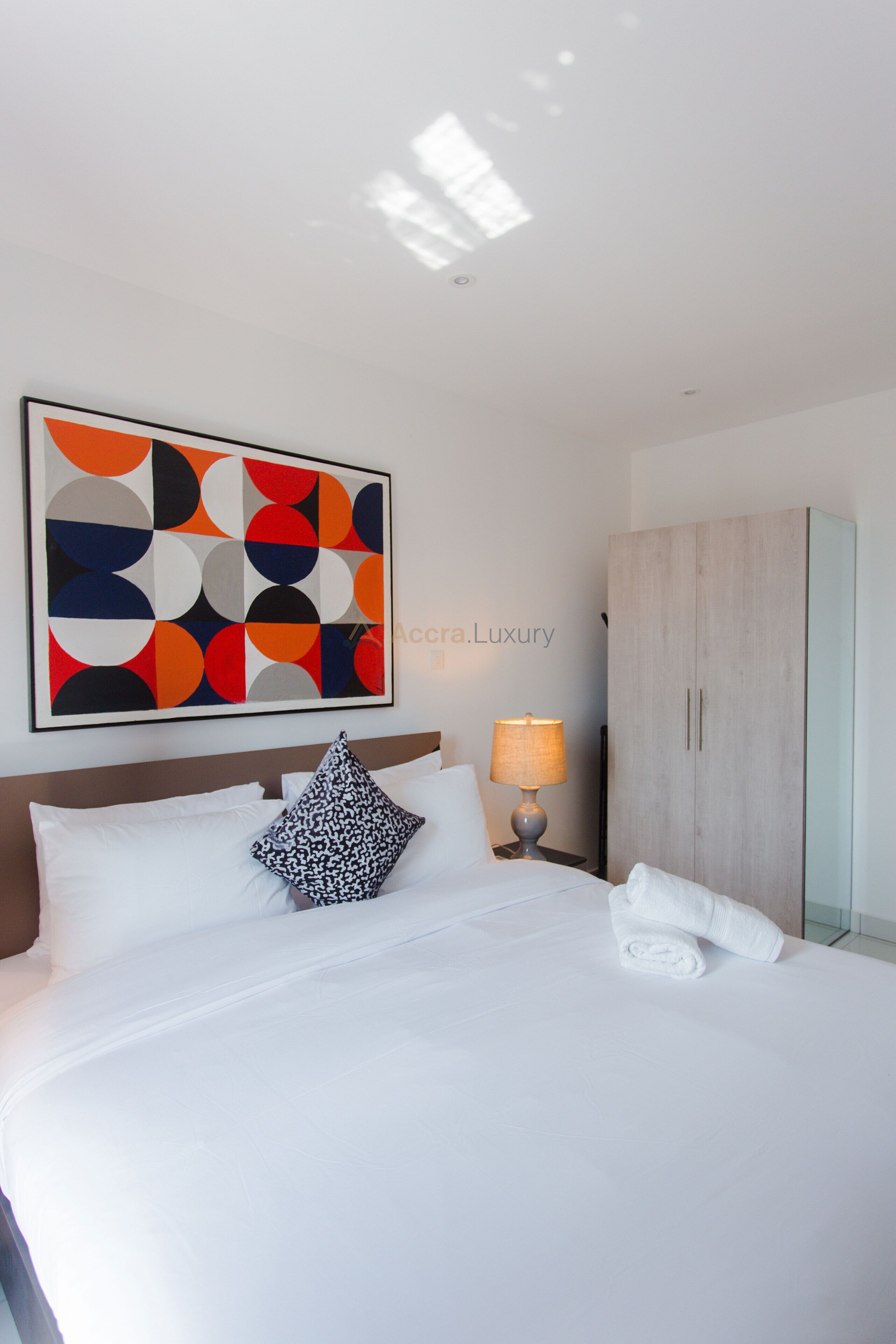 Une chambre d’hôtel avec un grand lit, une table de chevet avec une lampe, une armoire et une peinture abstraite aux couleurs vives accrochée au mur.