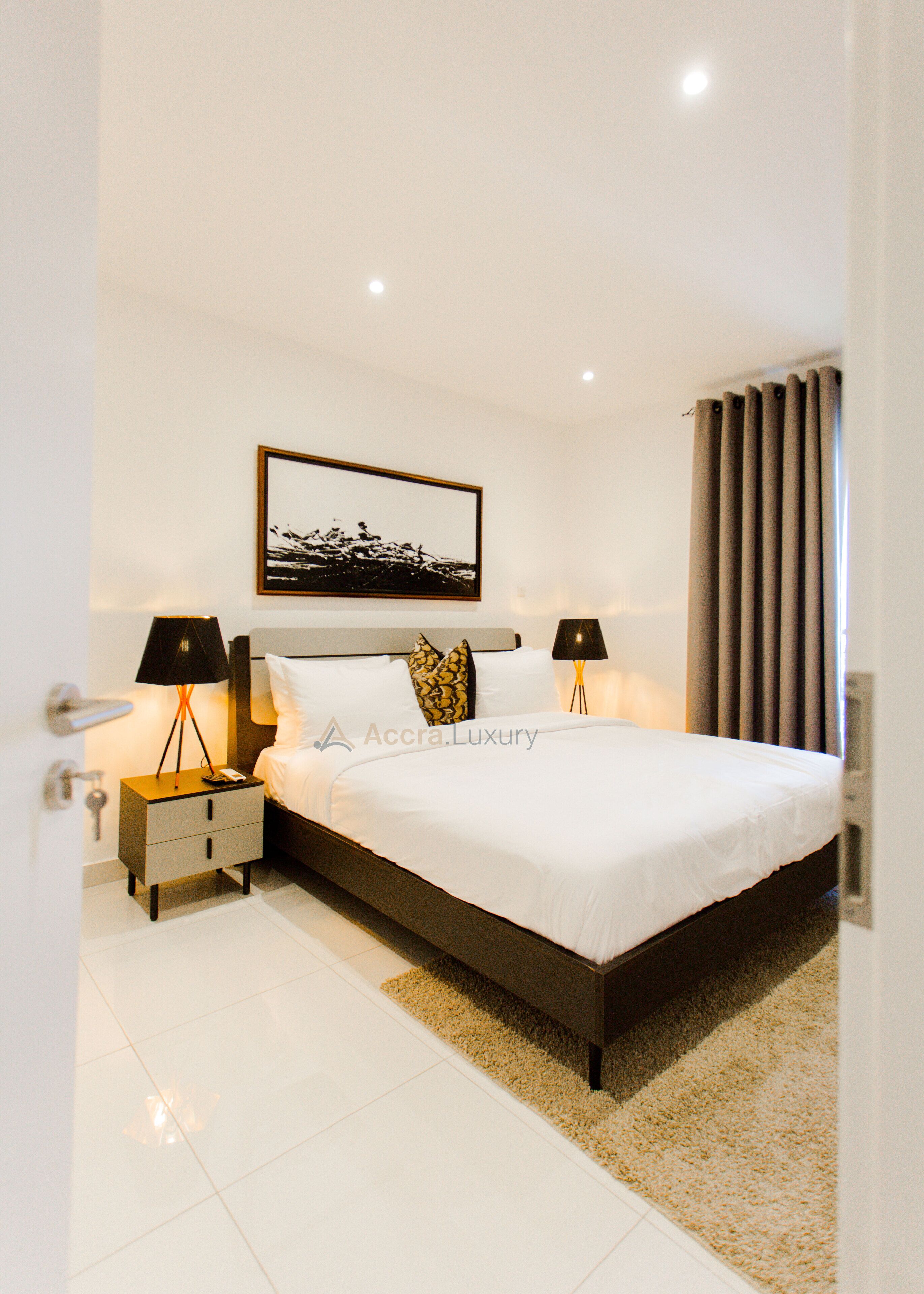 A modern bedroom with a large bed, two bedside tables with lamps, a framed picture on the wall, and a window with curtains.
