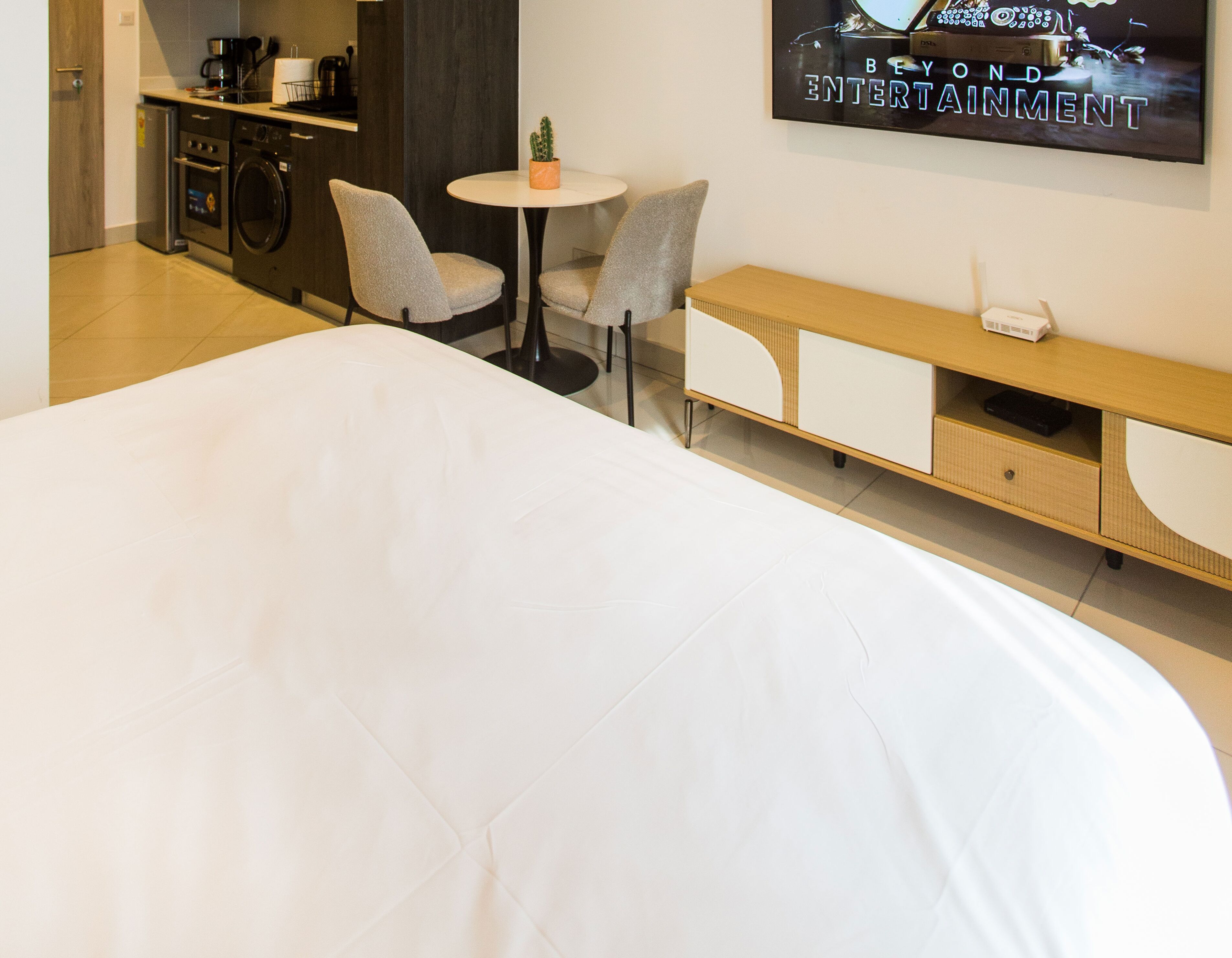 Quarto de hotel com uma cama grande, uma mesa redonda pequena com duas cadeiras, uma TV fixada na parede, um banco de madeira e uma máquina de lavar roupa.