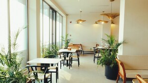 A spacious indoor dining area with large windows, multiple tables and chairs, and several potted plants.