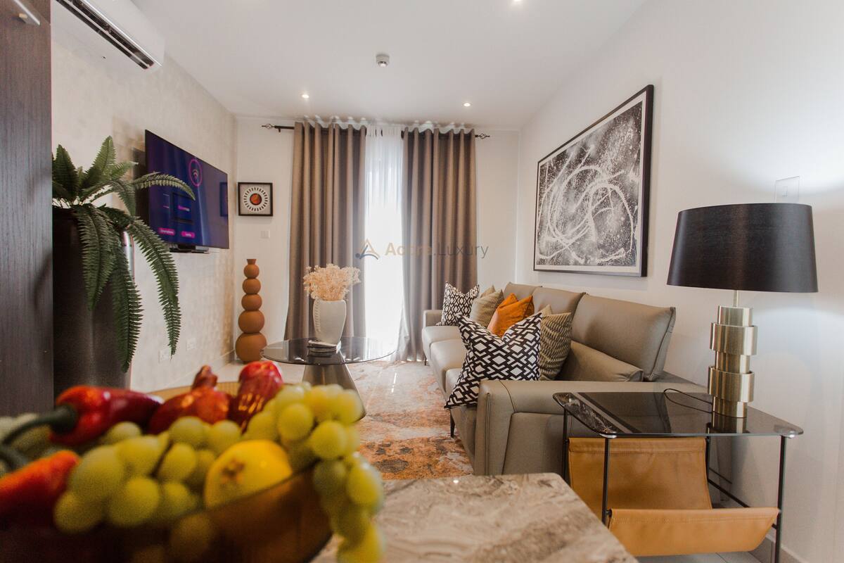 Un salón moderno con un sofá, una mesa de centro y un televisor. Hay una planta, un jarrón con flores y un frutero sobre la mesa.