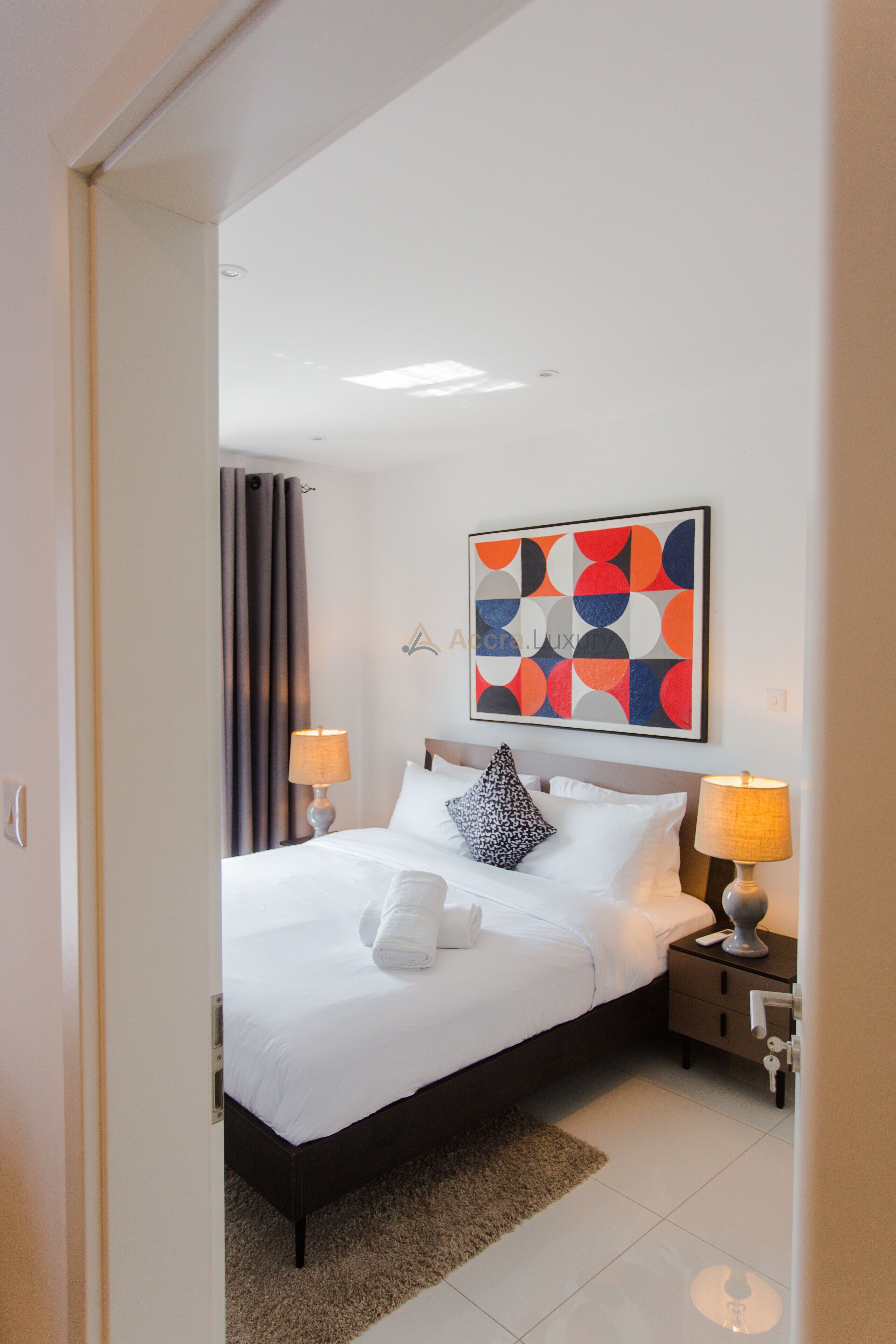 A modern hotel room with a neatly made bed, two bedside tables with lamps, a large abstract painting on the wall, and a view of the city through the window.