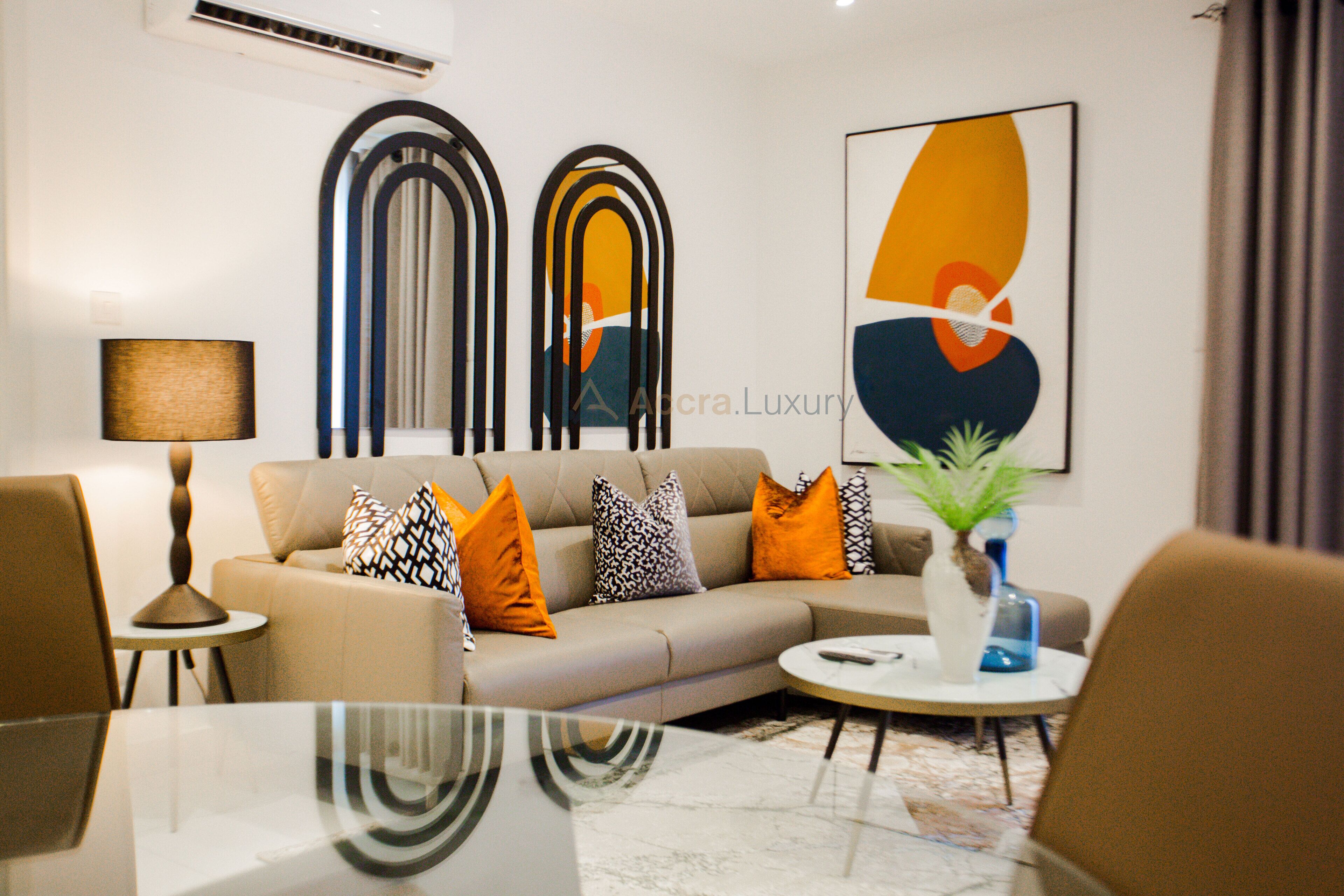 A modern living room with a sofa, coffee table, and decorative pillows. There is a lamp, a vase with a plant, and abstract wall art.