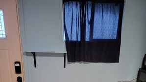 Eine kleine Küche mit Holztisch und zwei Hocker, einem Fenster mit Vorhängen sowie einem eingebauten Backofen.