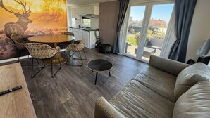 A modern living room with a sofa, a dining table with chairs, a kitchen area, and a large window with curtains.