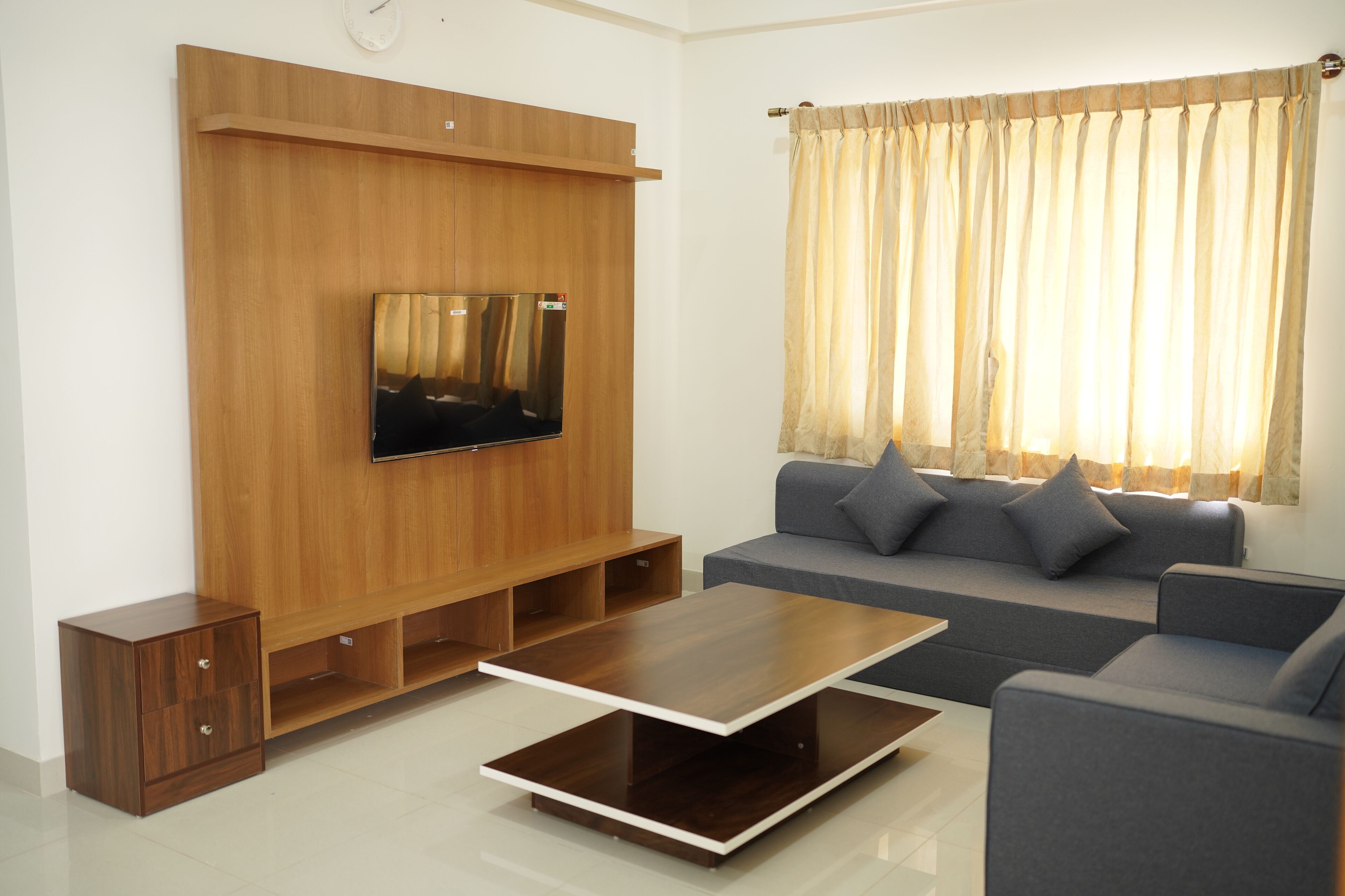 A modern living room with a flat-screen TV mounted on a wooden wall, a grey sofa with cushions, a wooden coffee table, and a window with curtains.